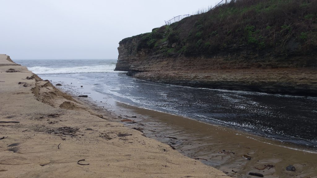 Sand bank from upper left edge down to bottom. Ocean in the background. Water flowing between ocean on the left and right edge of photo. Cliff occupying top right corner of photo.